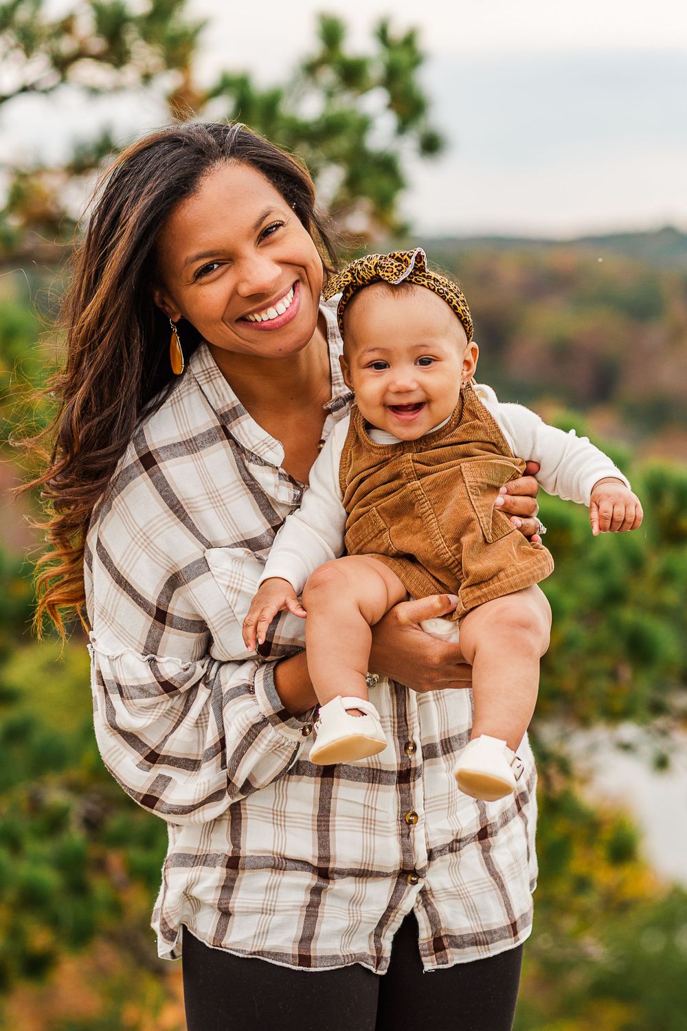 A joyful autumn outdoor portrait showing a parent in plaid holding a baby wearing brown corduroy against a nature backdrop.
