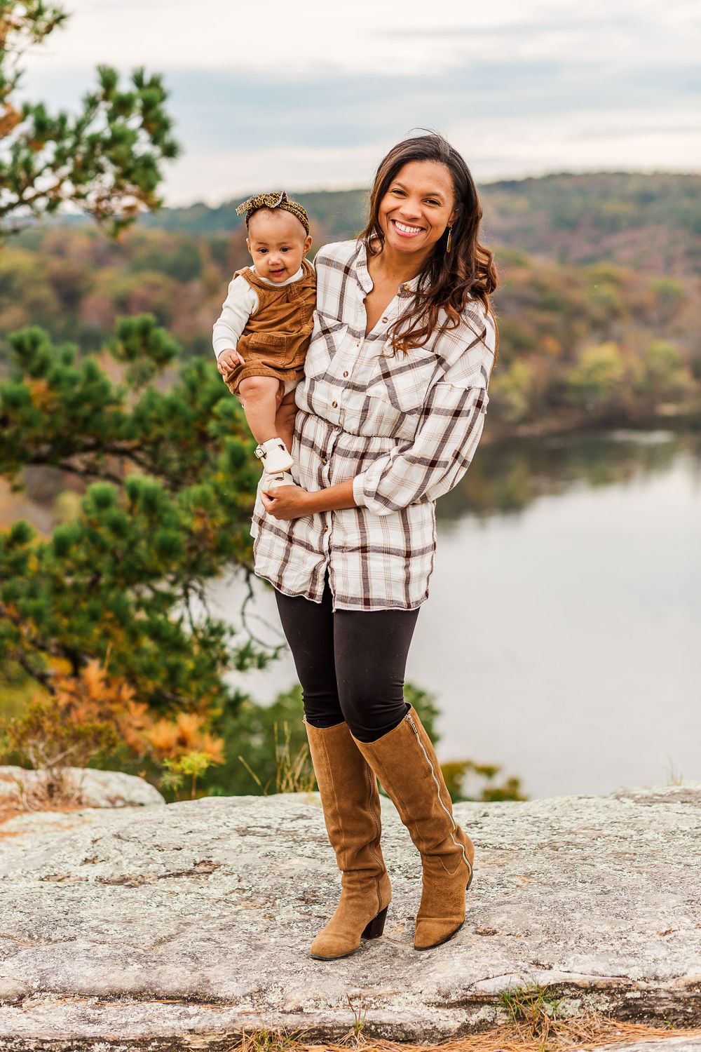 Outdoor autumn portrait with parent holding baby while standing on rocky overlook with water view.