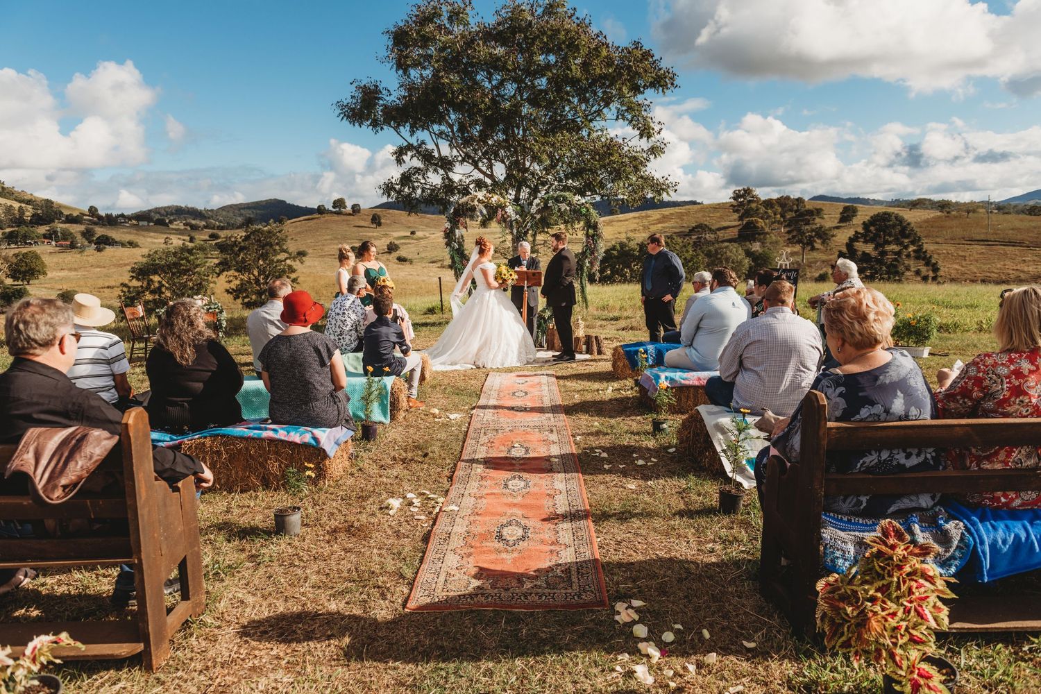 Outdoor wedding ceremony setup with vintage rugs and chairs in a rural landscape.