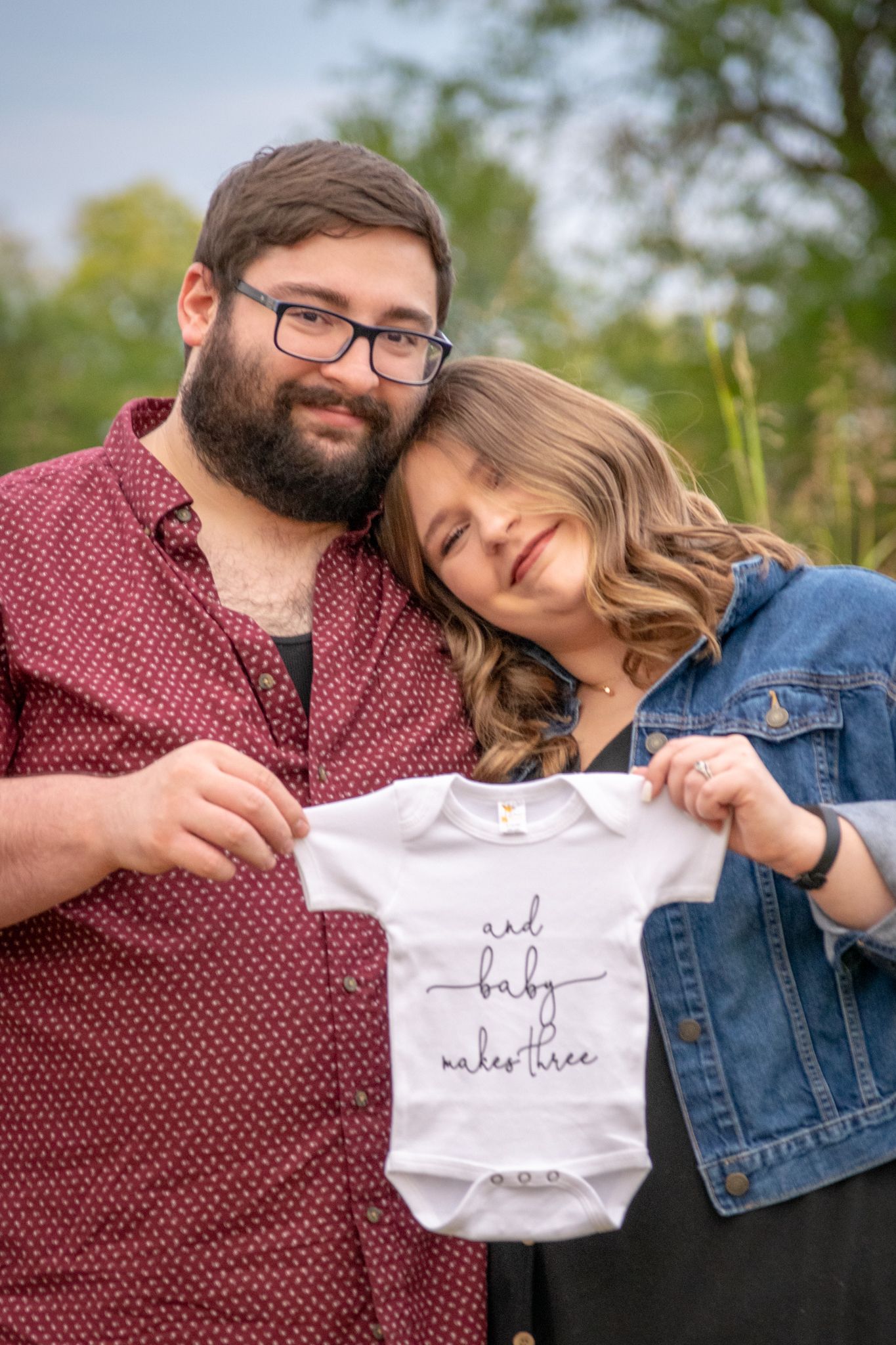 A couple snuggles holding a onesie that reads "And baby makes three" Pregnancy announcement session. Kansas City, MO