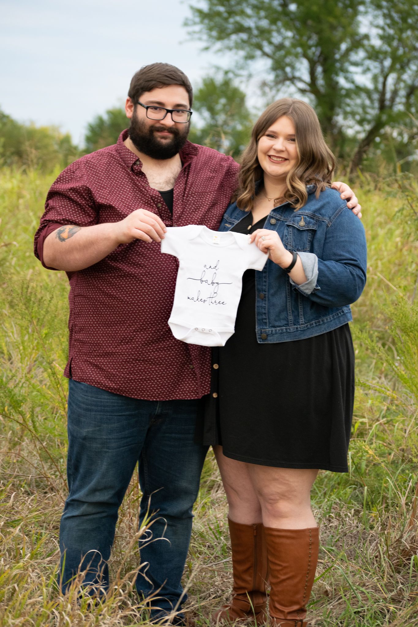 Expecting parents smile and hold a onesie that reads "And baby makes three" Baby announcement session. Kansas City MO