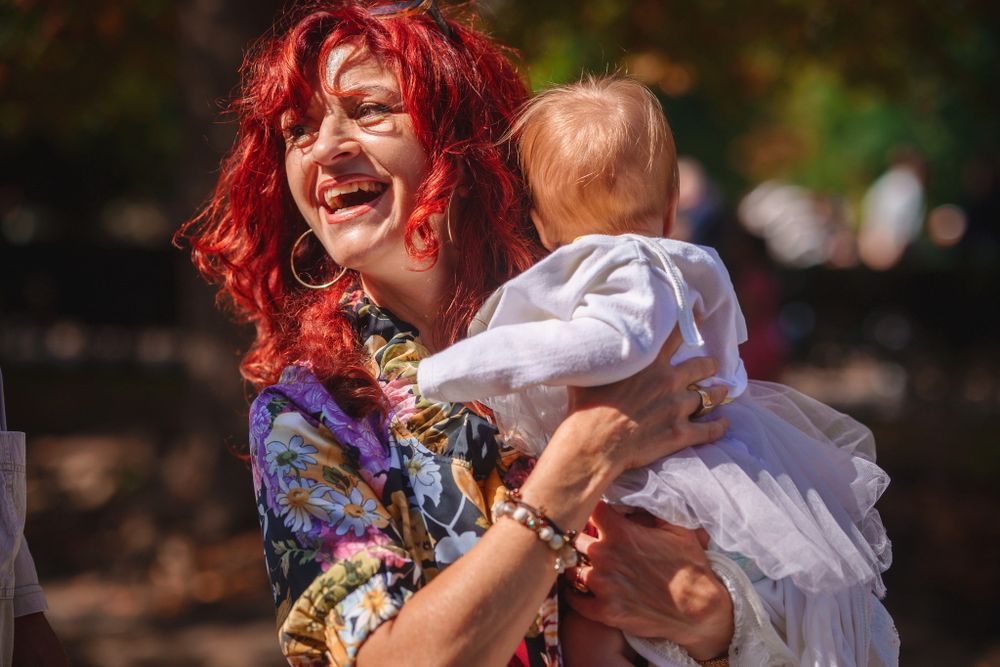A woman with bright red curly hair laughs while holding a small baby outdoors.