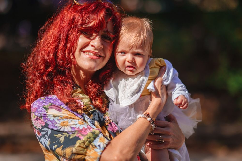 A woman with vibrant red hair holds a baby wearing white in an outdoor setting.