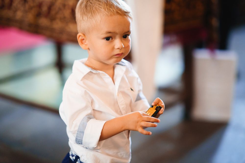 Small toddler in white shirt and blue shorts holds a snack while standing indoors.