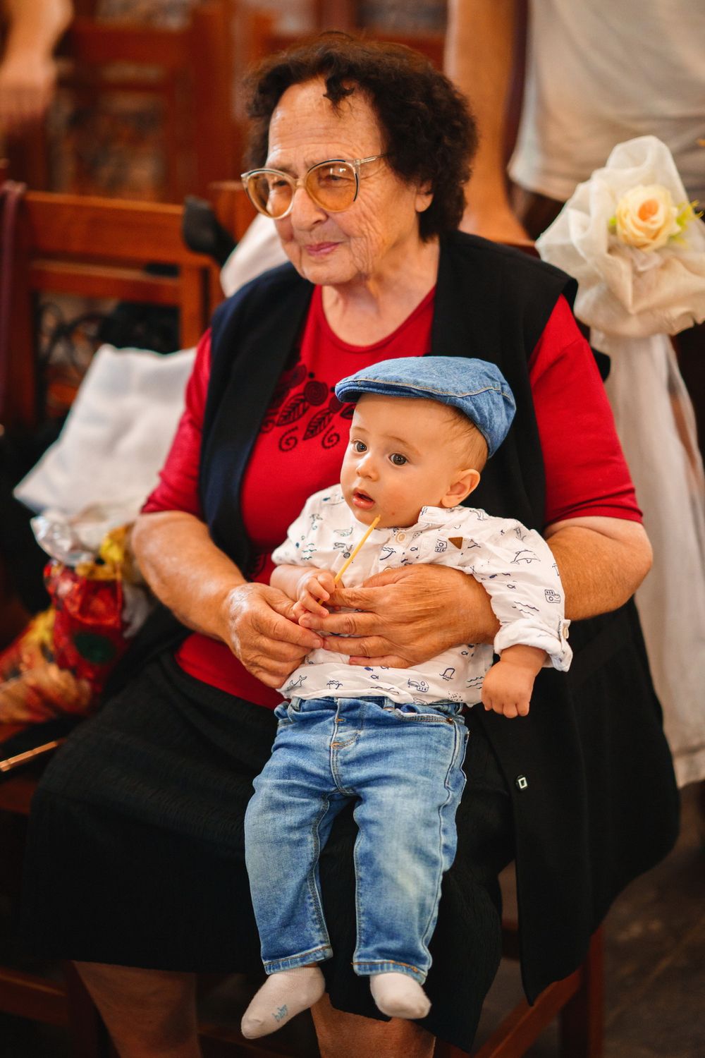 Senior woman in red sweater holds baby wearing jeans on her lap in church setting.