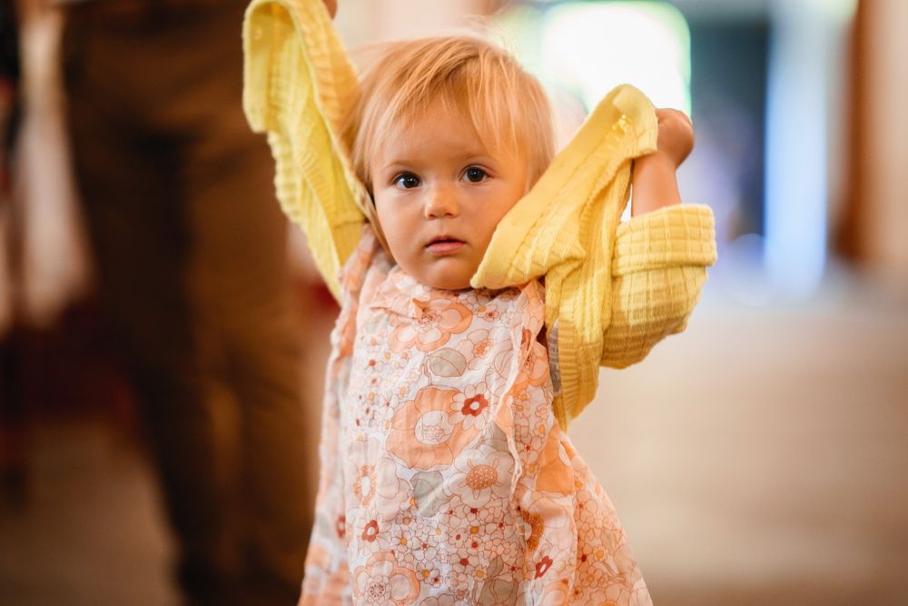 Baby in floral dress holds yellow cloth while standing indoors.
