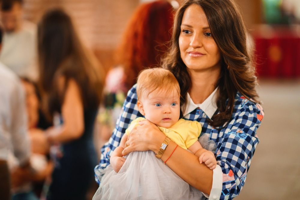 Woman in plaid shirt holds infant wearing white dress at indoor gathering.