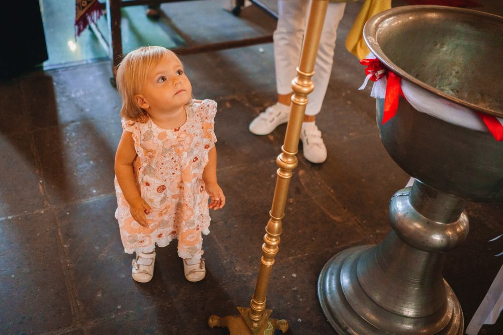 Small child in a white lace dress standing next to an ornate brass candlestick in a church.
