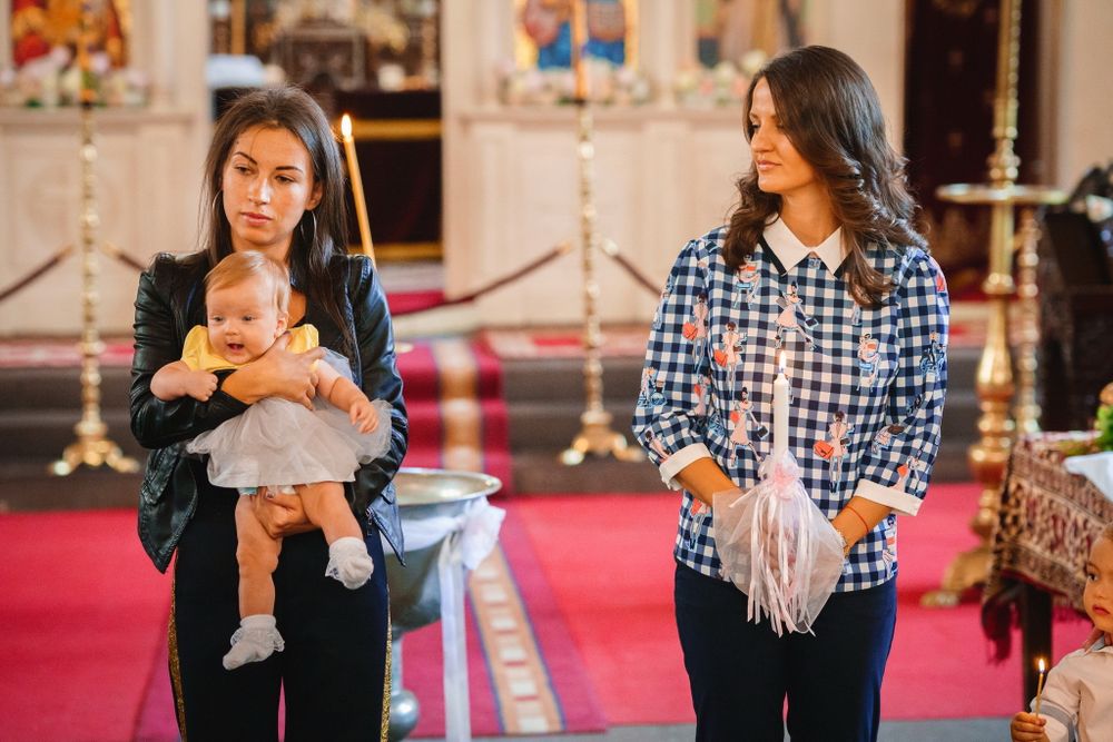 A young baby and two women stand inside a historic church with red carpeting and ornate architecture.