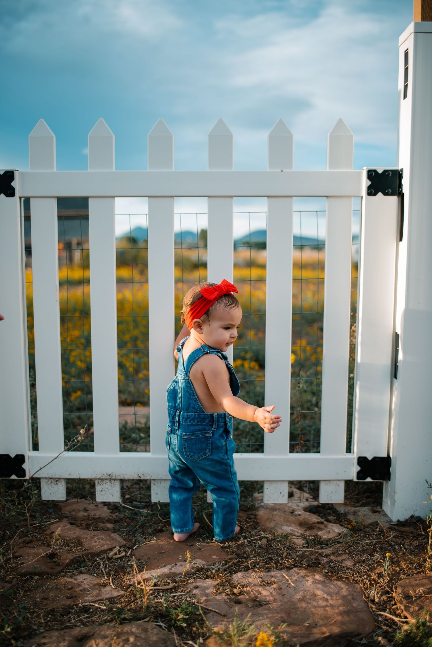Small child in overalls standing by white picket fence gate.