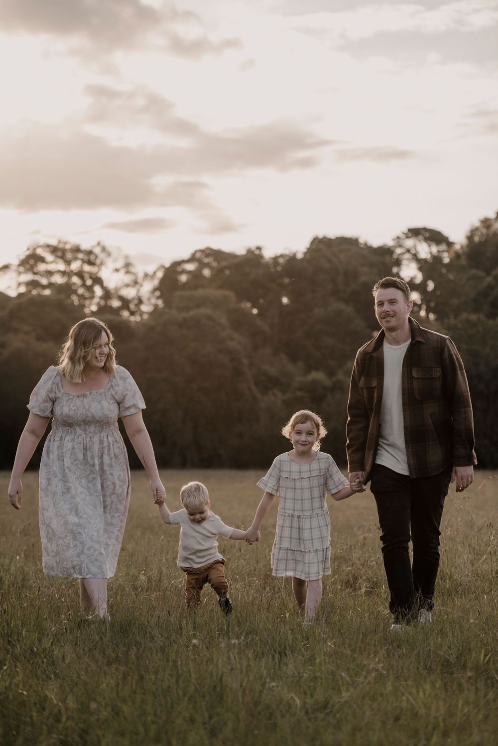 A family walks together holding hands through a grassy meadow at sunset in casual attire.
