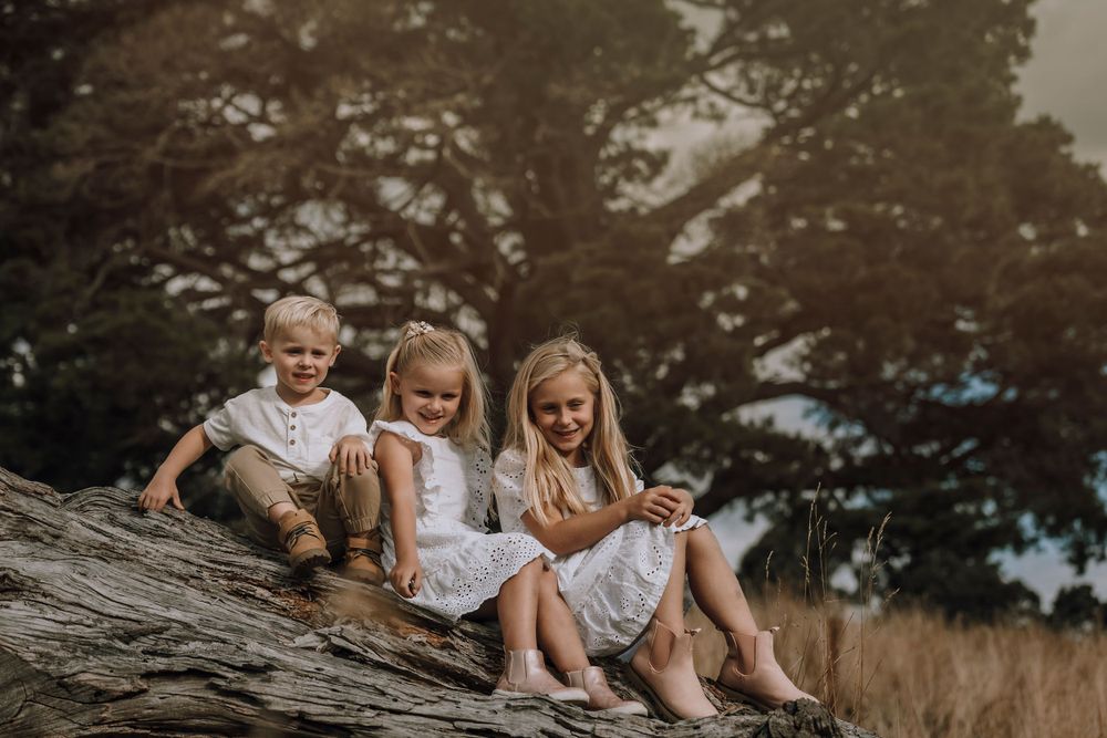 Three children in white clothing sit together on a large rock in a natural outdoor setting during golden hour.