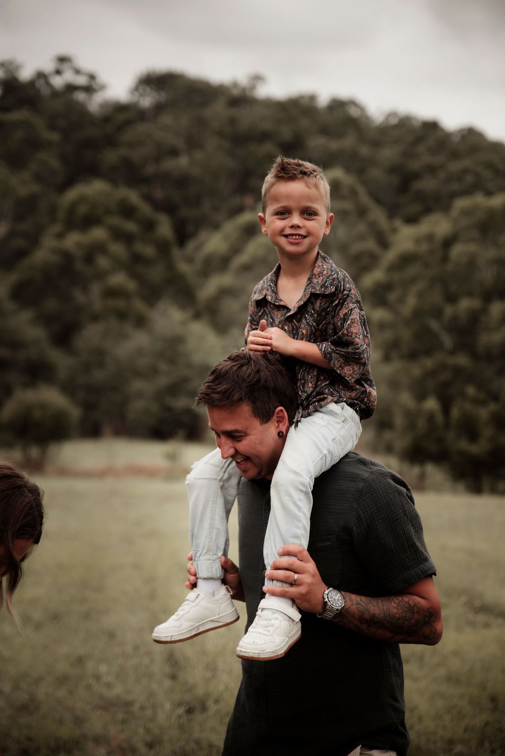 A child sits on someone's shoulders in a grassy field with trees in the background at dusk.