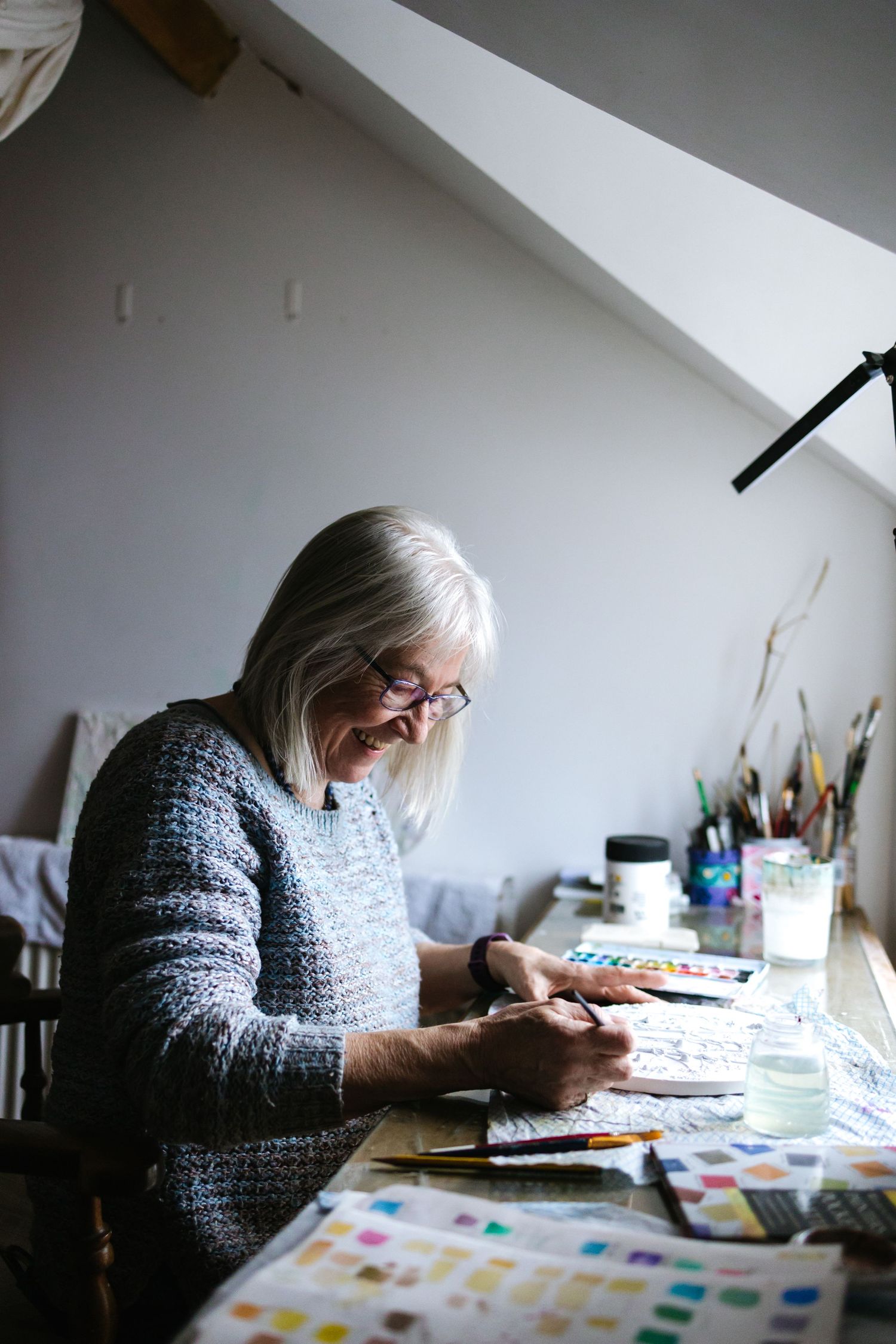 Artist in cozy sweater working on colorful artwork at desk near window with art supplies spread around.