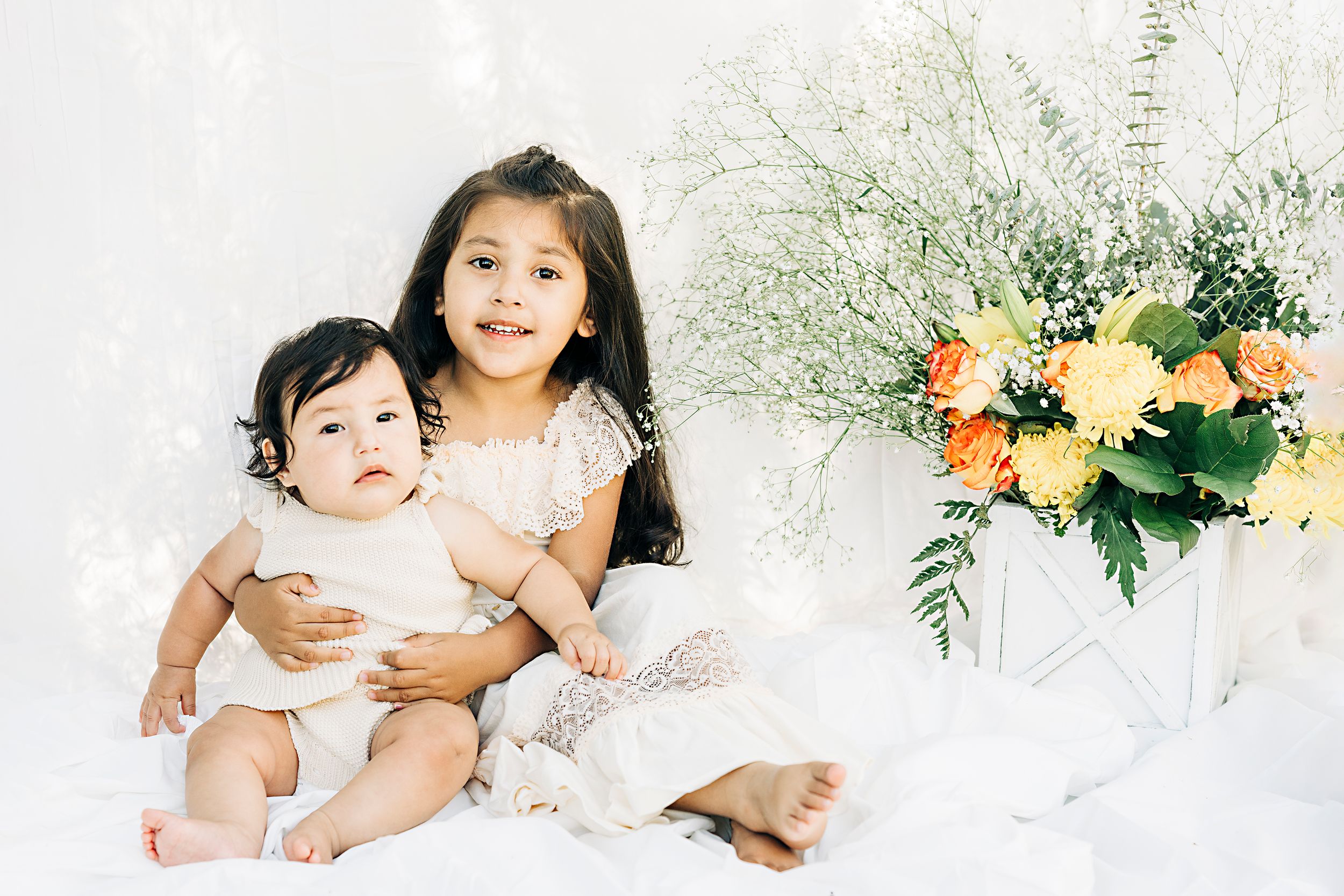 Two children in white outfits sitting together with floral arrangement on white backdrop.