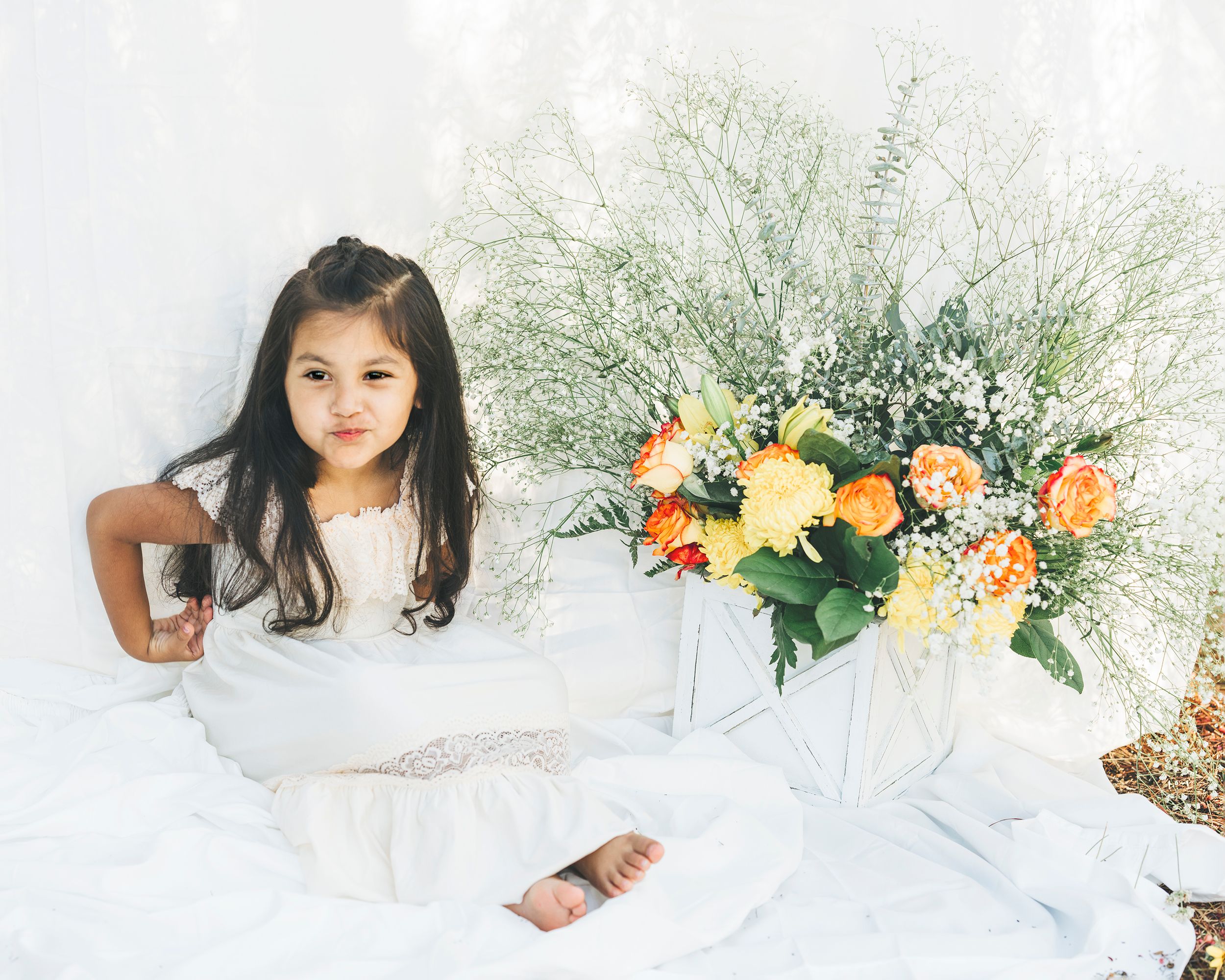 Child in white dress posing next to orange and yellow floral arrangement on white backdrop.