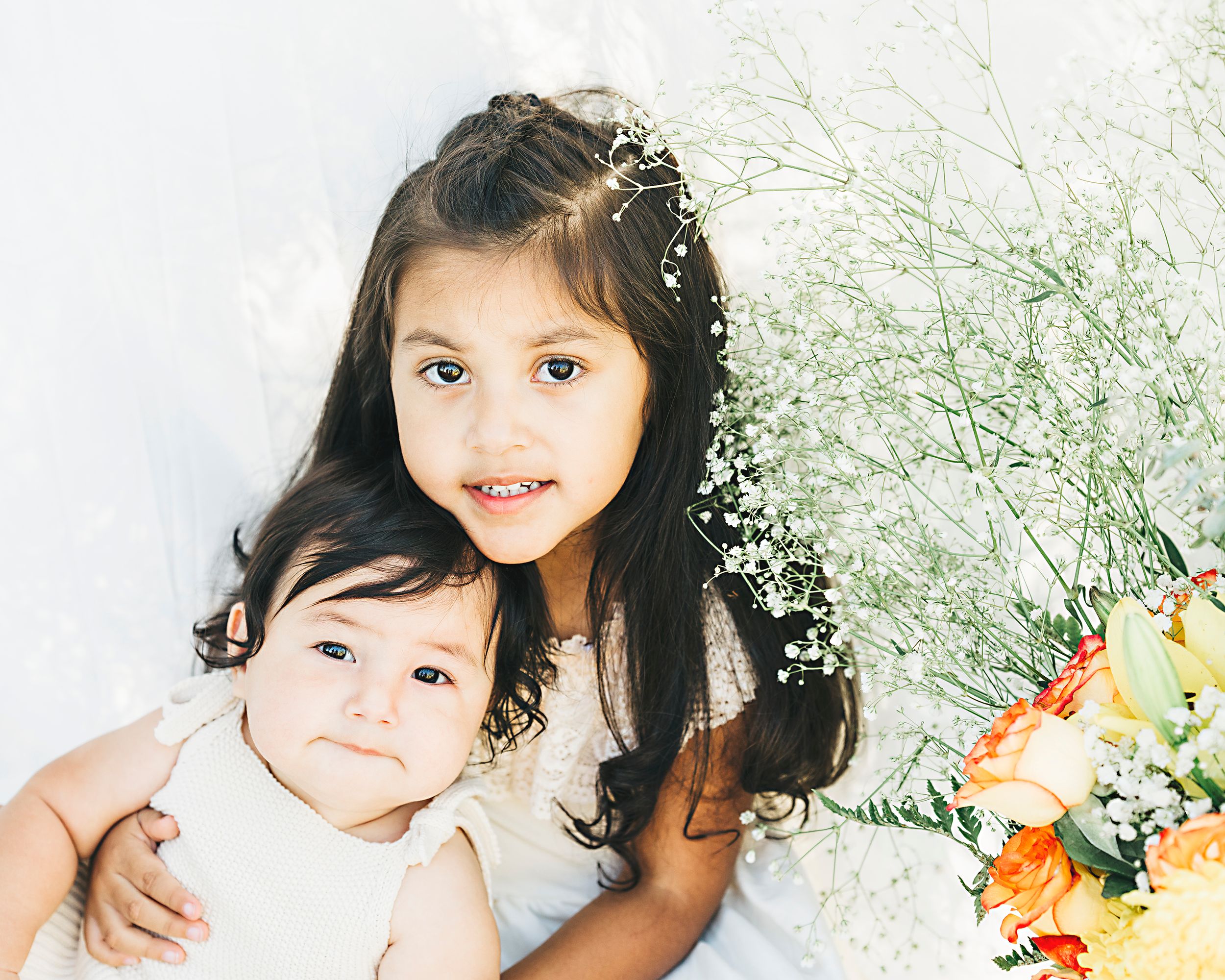 Two siblings posing together with orange and yellow floral arrangement against white background.