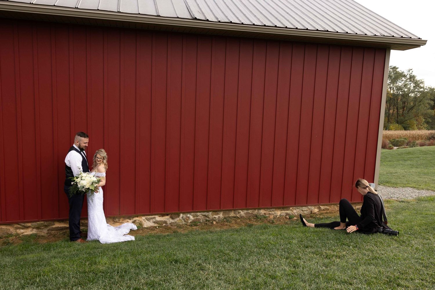 A photographer crouches to capture a wedding moment against a red barn wall.