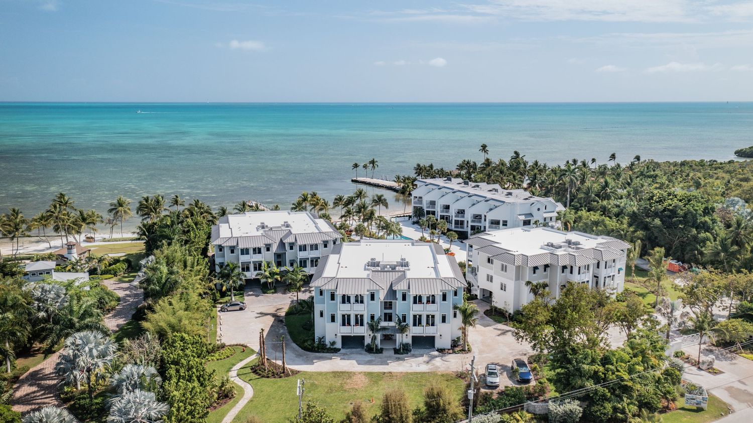 Aerial view of luxury white beachfront condos surrounded by palm trees along turquoise waters.