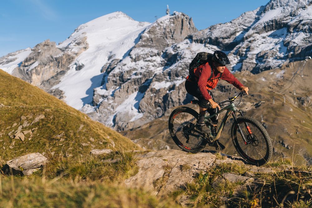 Mountain biker jumps over terrain with snow-covered alpine peaks in the background.