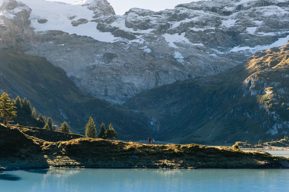 Snow-capped mountain peaks reflect in turquoise alpine lake with golden light on hillside.