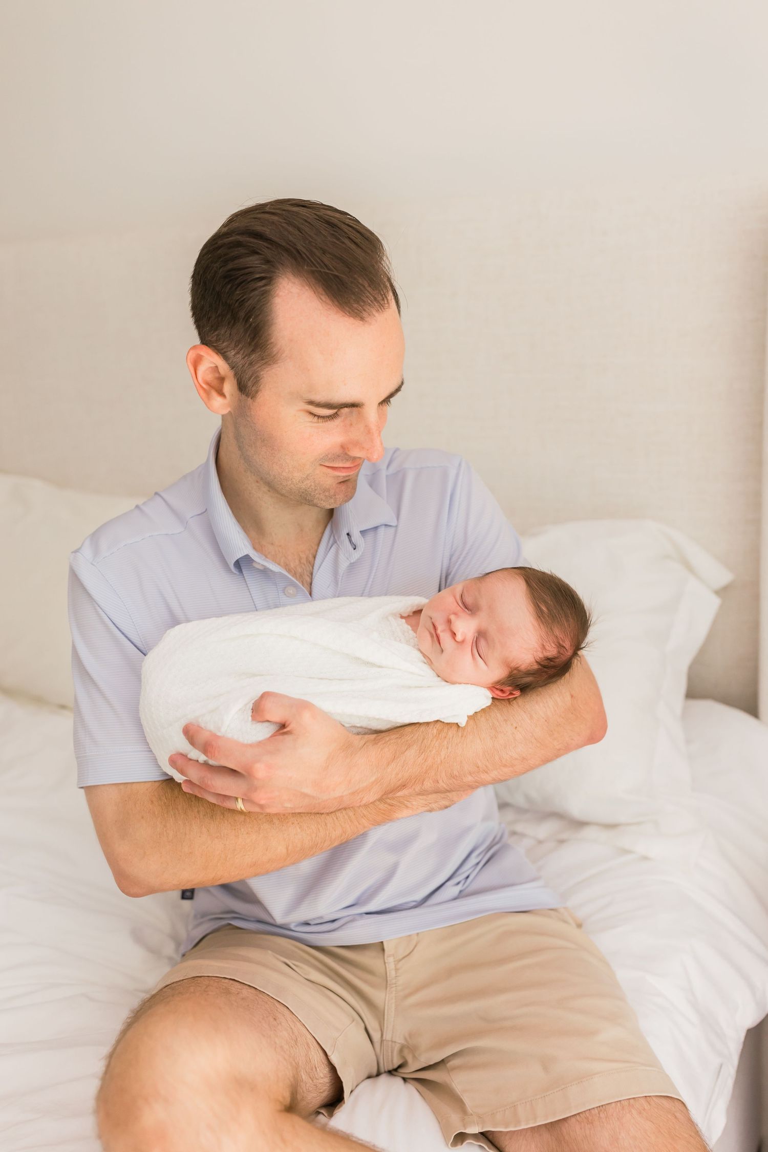 A father in light blue shirt tenderly holds his sleeping newborn wrapped in white.