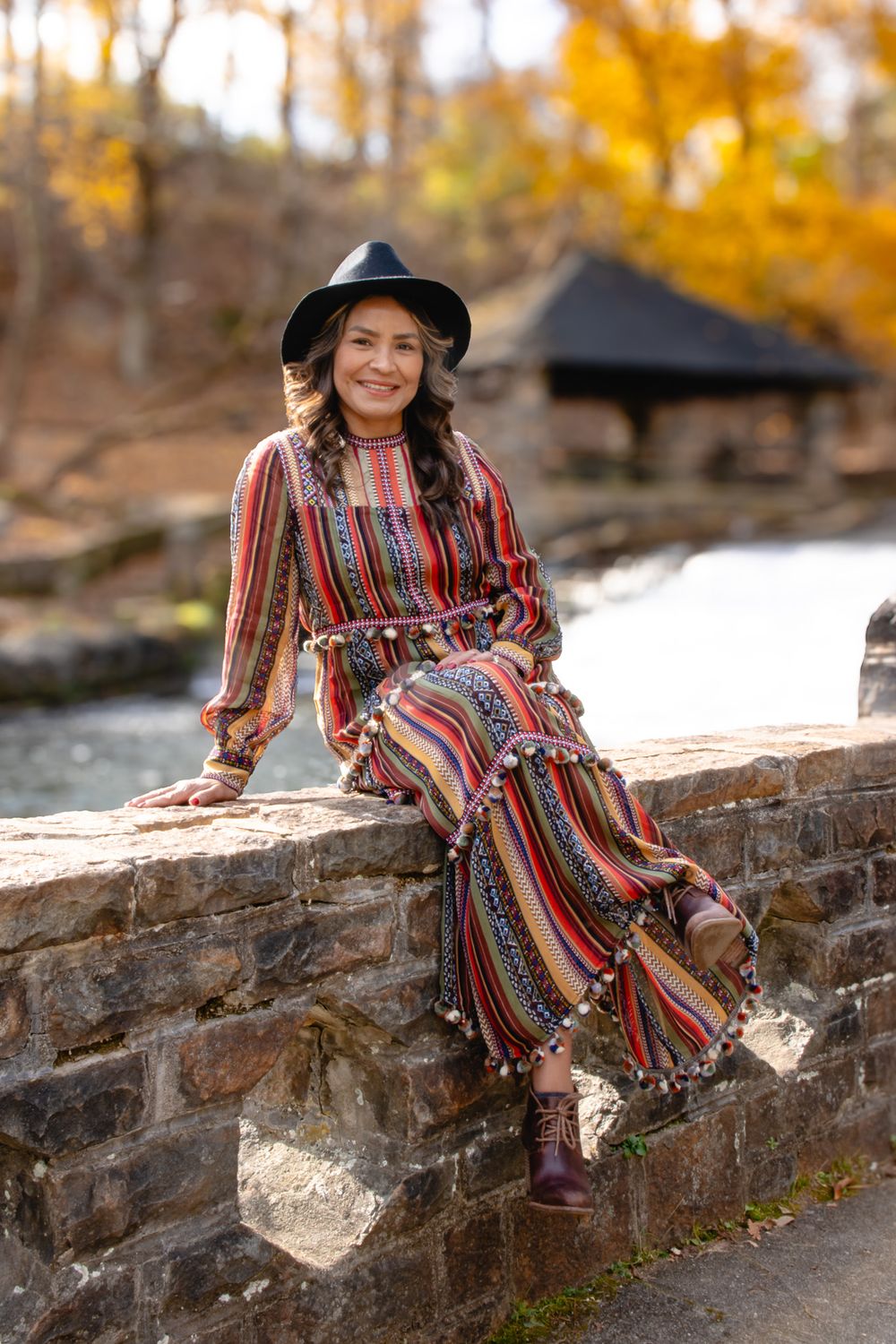 Woman in colorful striped boho dress and black hat sits smiling on stone wall with fall foliage behind her.