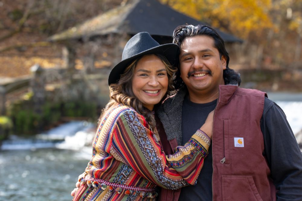 A smiling couple poses together outdoors, the woman wearing a colorful wrap and the man in a vest and black hat.