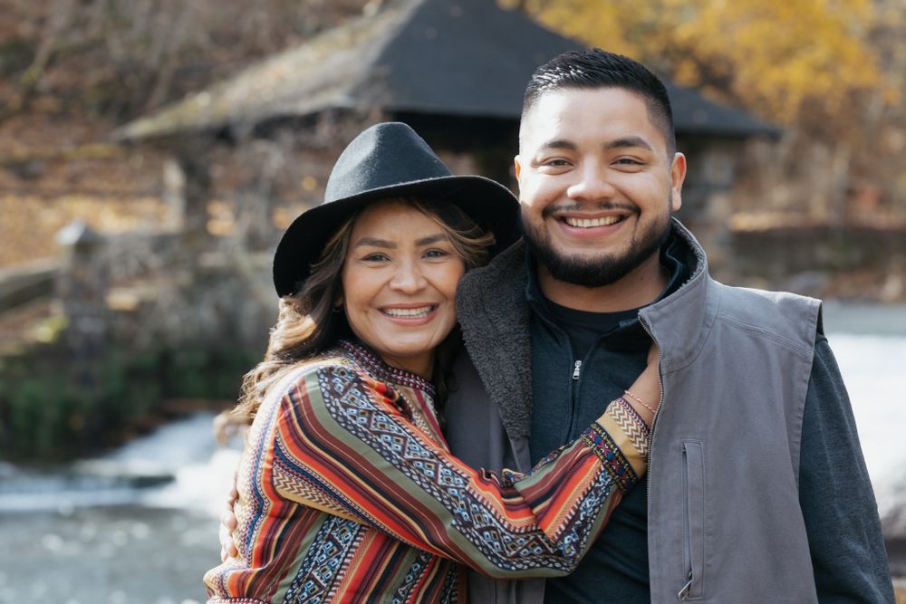 Woman in colorful boho wrap and black hat smiling with young man in gray jacket outdoors in fall.