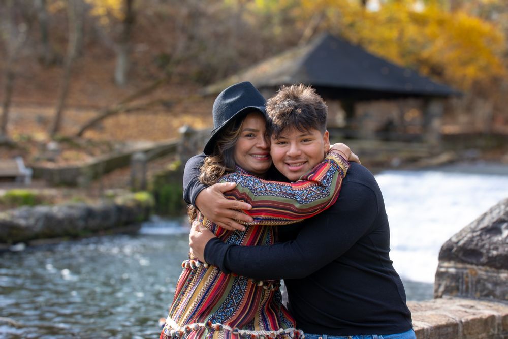 Two people sharing a warm embrace and smiling outdoors near a pond with autumn trees in background.
