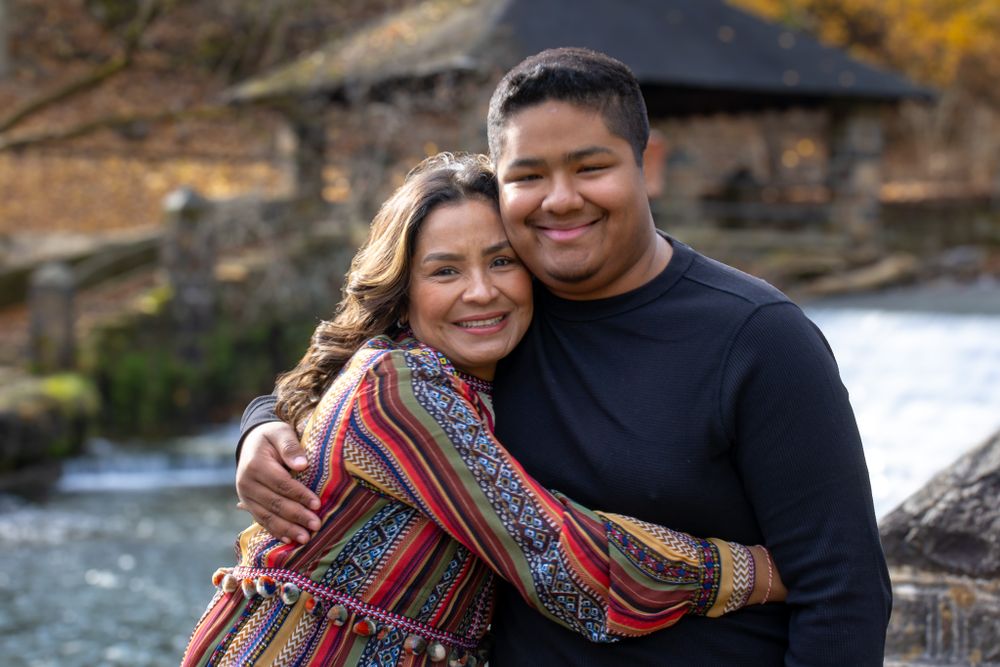 A woman and young man share a warm embrace outdoors near a river with autumn foliage in the background.