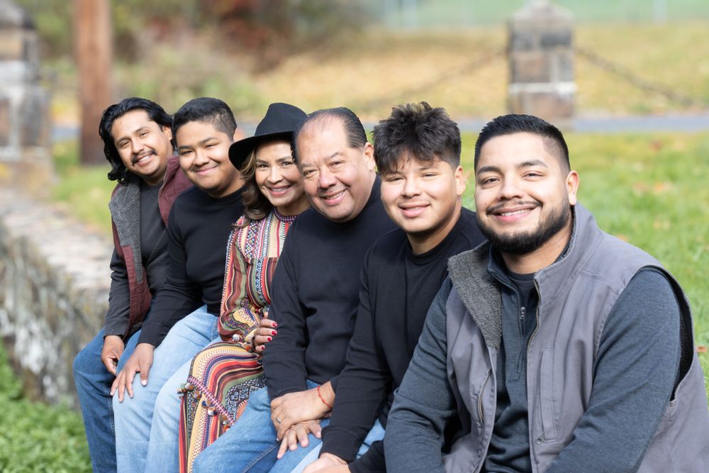 Latino family of six sitting in a row outdoors, smiling warmly, with woman in colorful dress and man in gray vest.