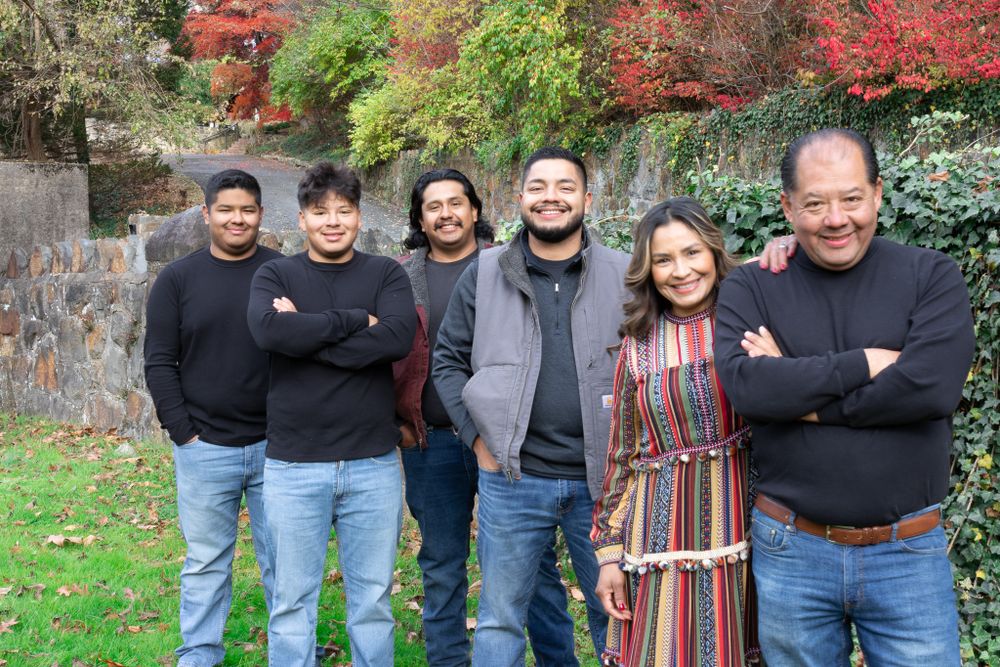 Group of six people in black sweaters and one woman in striped dress posing outdoors in autumn garden.