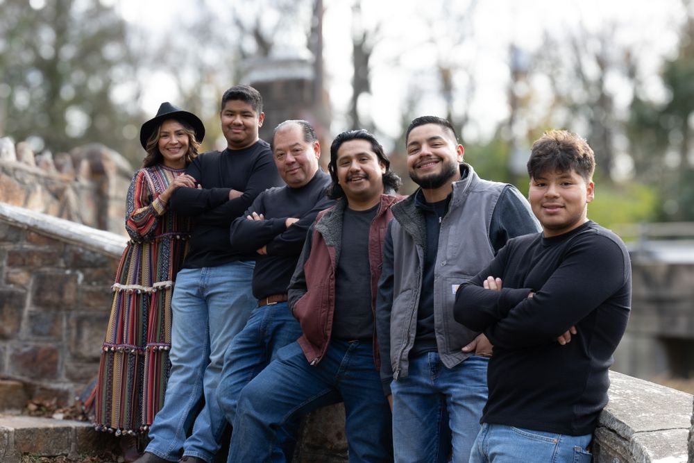 Latino family of six posing outdoors on stone steps, smiling at camera in casual dark clothing in a park setting.