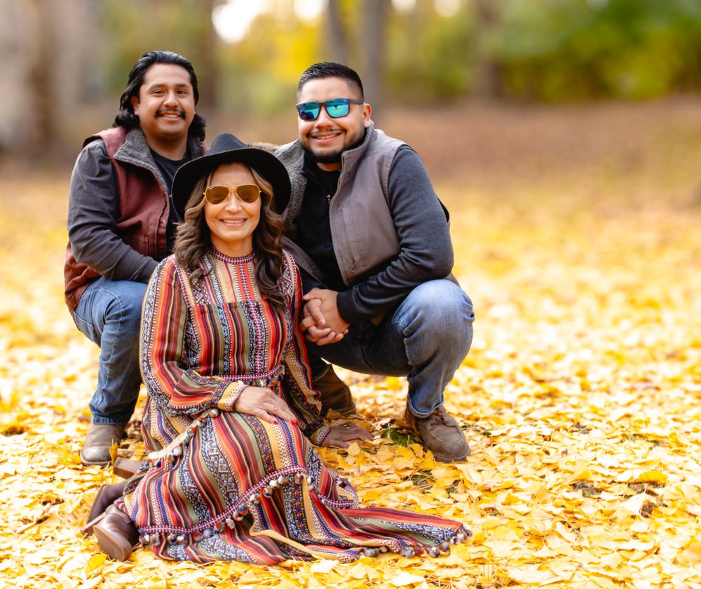 Three people pose together in autumn leaves, woman in colorful dress and black hat seated between two men.