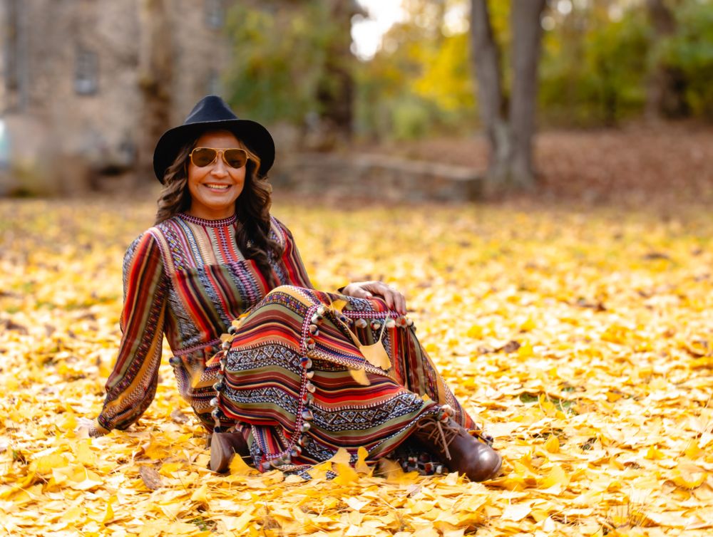 Woman in colorful striped dress and black hat sitting and smiling on golden autumn fallen leaves.