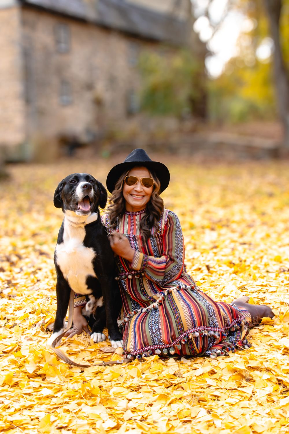 Woman in colorful striped dress and black hat sits with large black and white dog on golden autumn leaves.