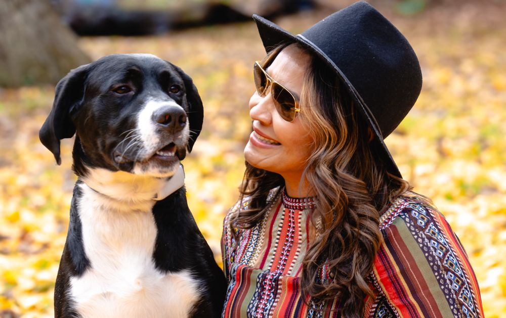 Smiling woman in a black hat and colorful top shares a joyful moment with a black and white dog outdoors.