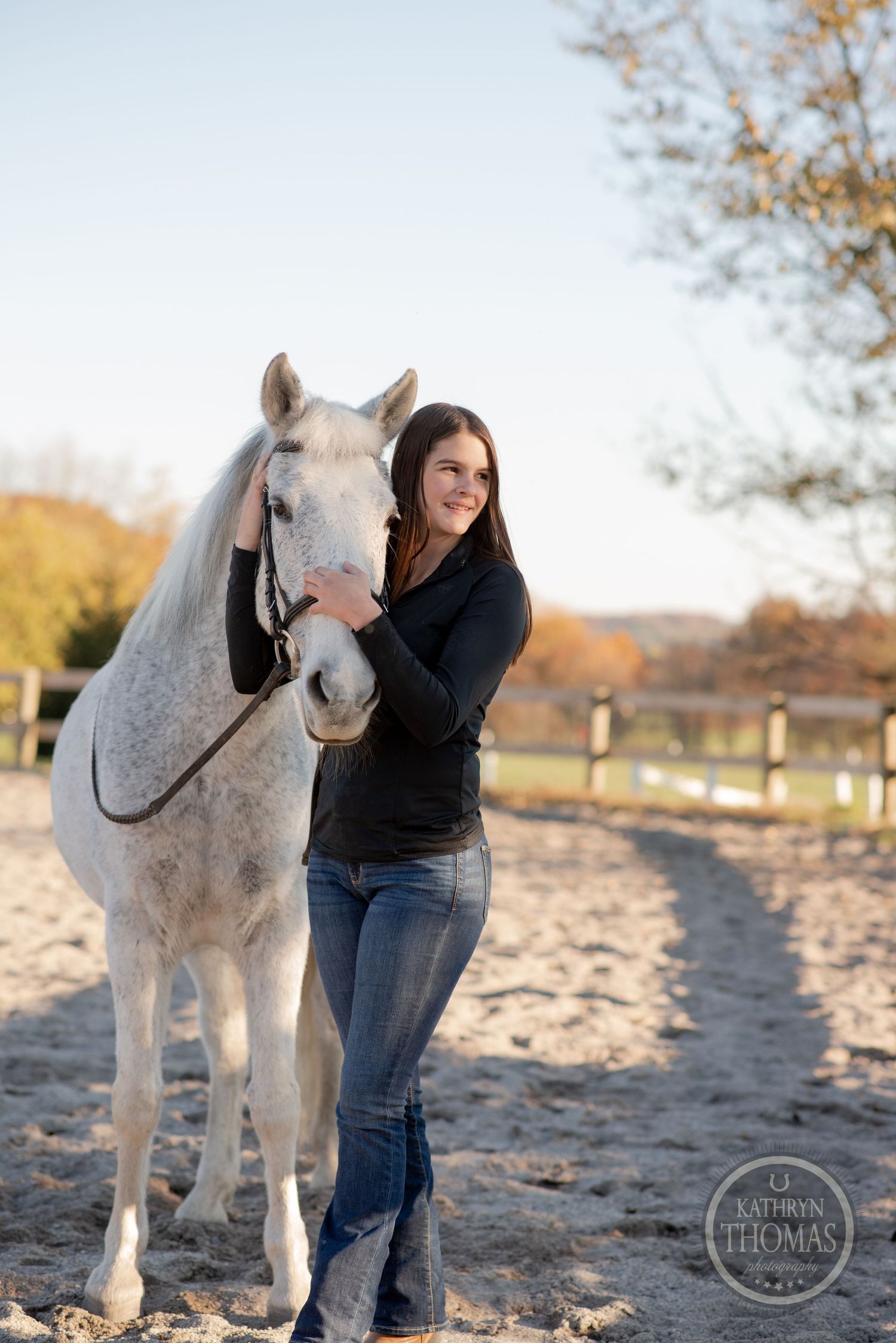 Person standing beside a white horse in a sandy arena with fall foliage in background.