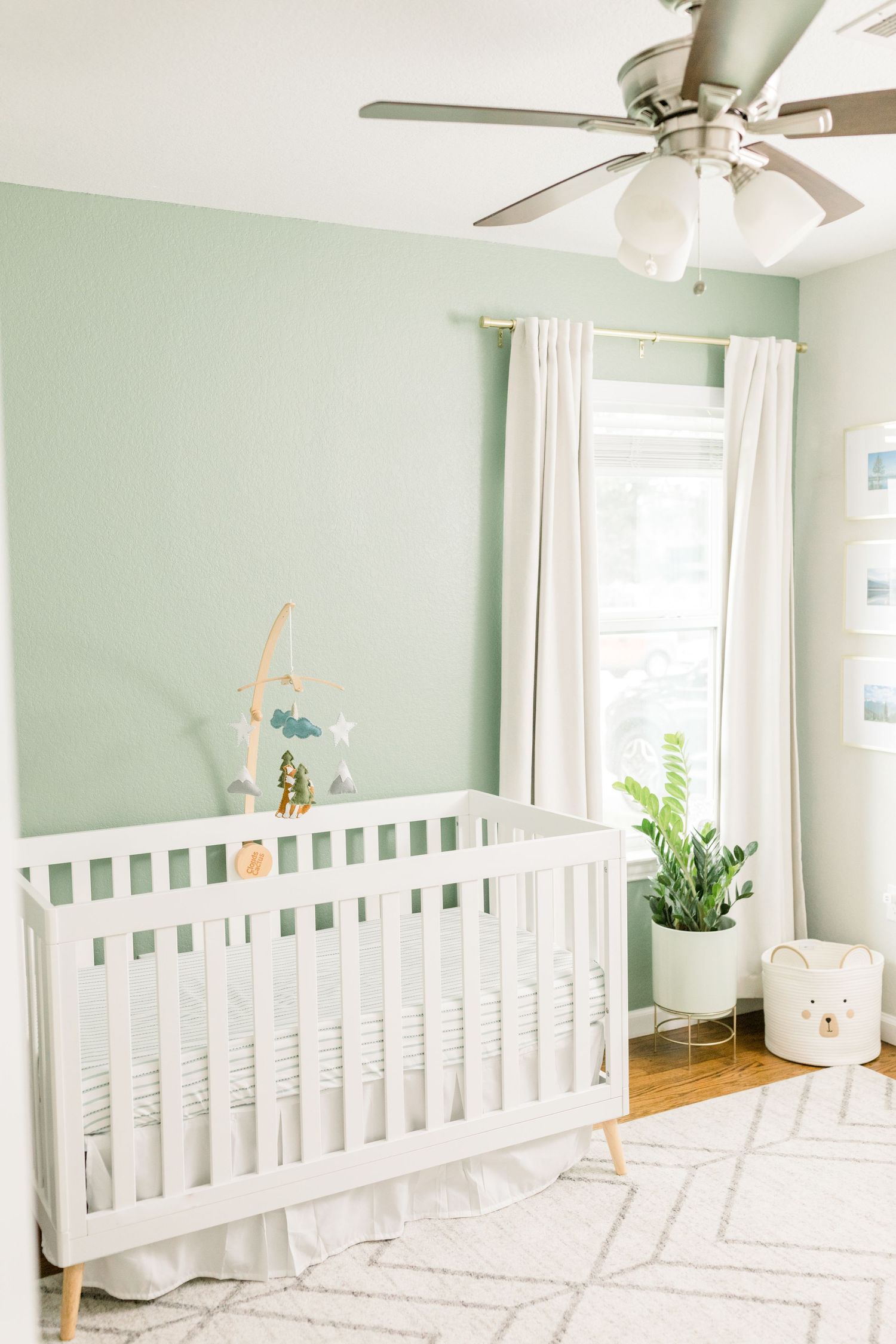 Serene nursery with mint green walls, white crib, ceiling fan and sheer curtains on a geometric patterned rug.