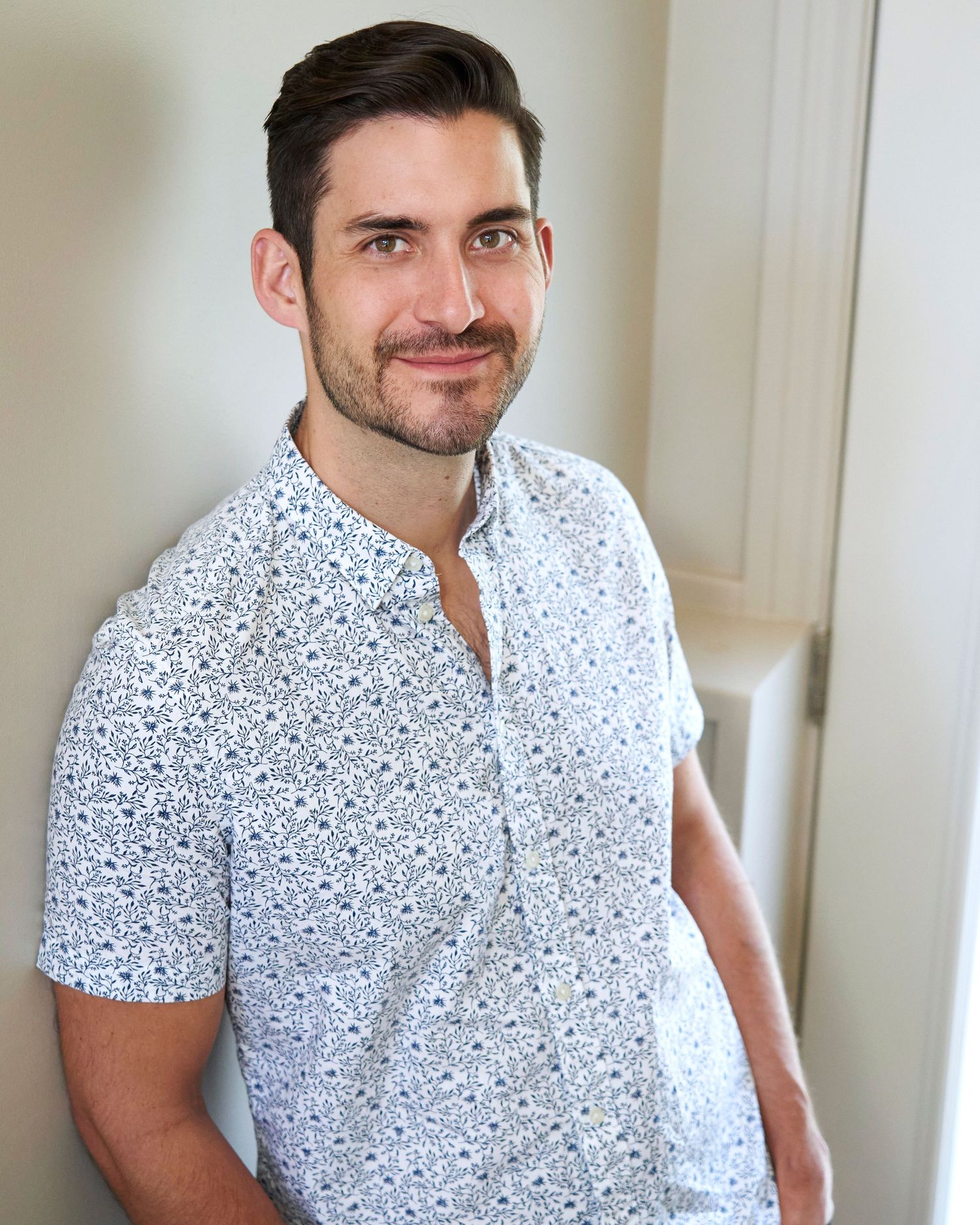 A person wearing a white patterned short-sleeve button-up shirt stands against a light-coloured wall creating a friendly headshot portrait.