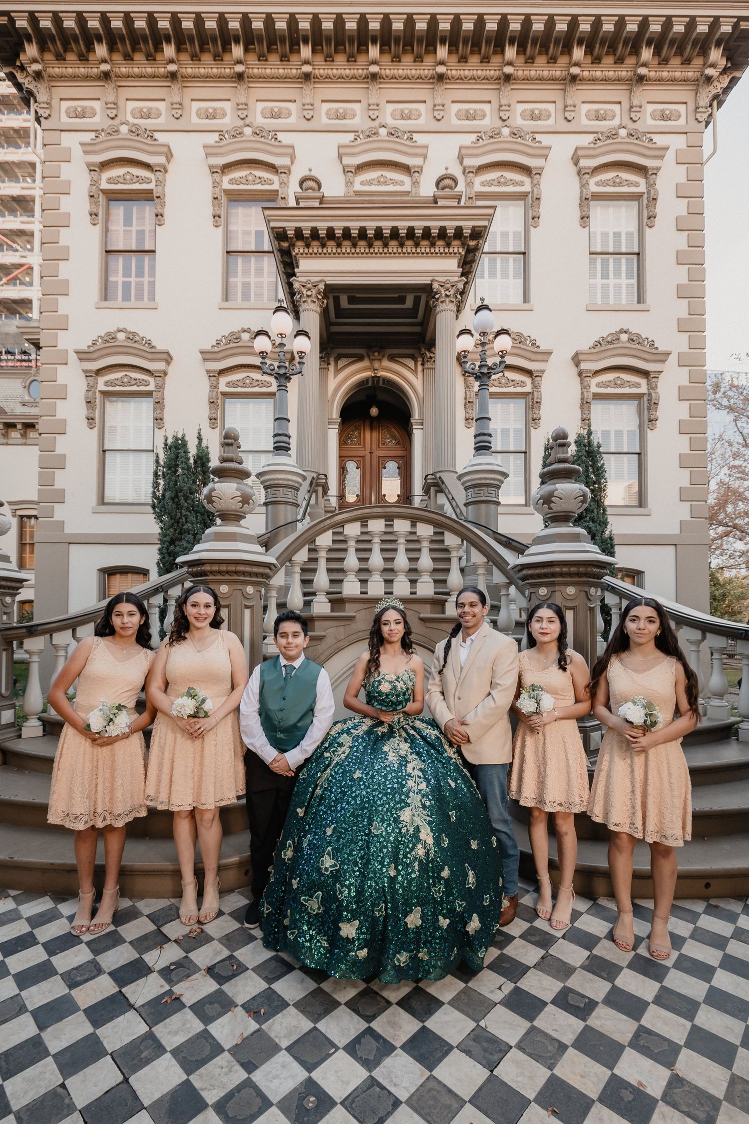 Quinceañera celebration group photo with attendants in peach dresses on mansion steps.