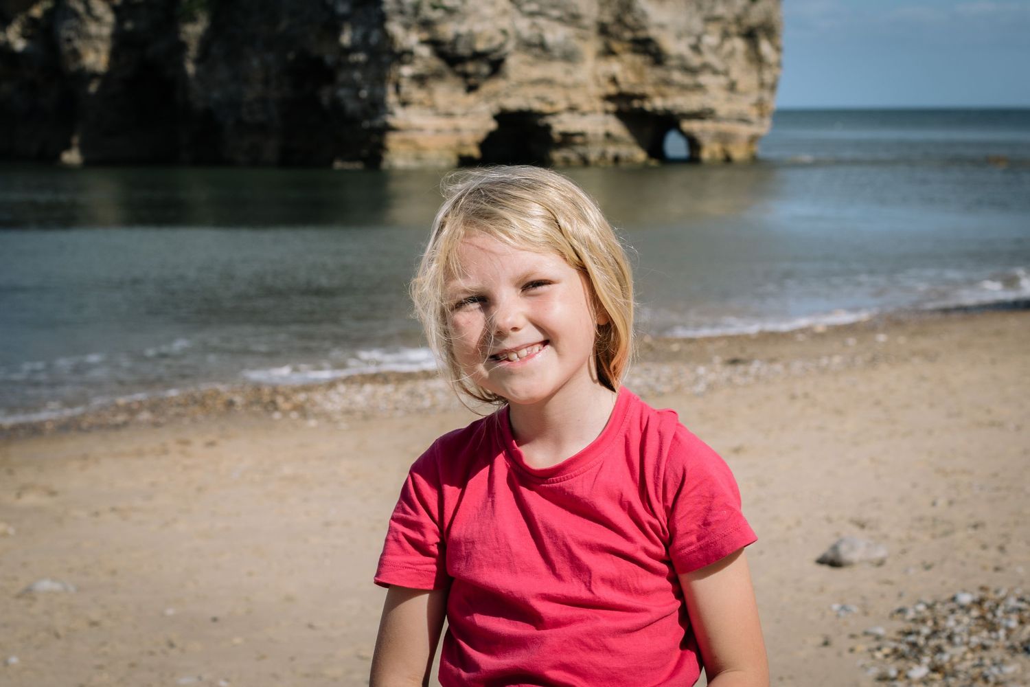 Child wearing a bright red t-shirt sitting on a beach with dramatic coastal cliffs in background.