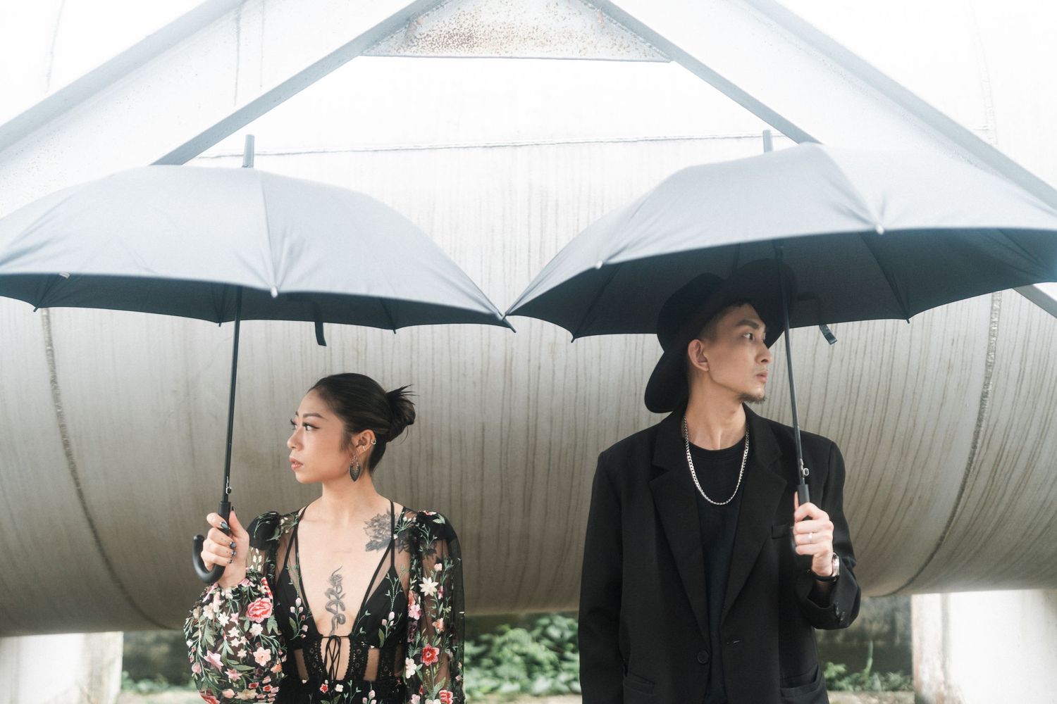 A stylish couple poses back-to-back holding black umbrellas against a white architectural backdrop.