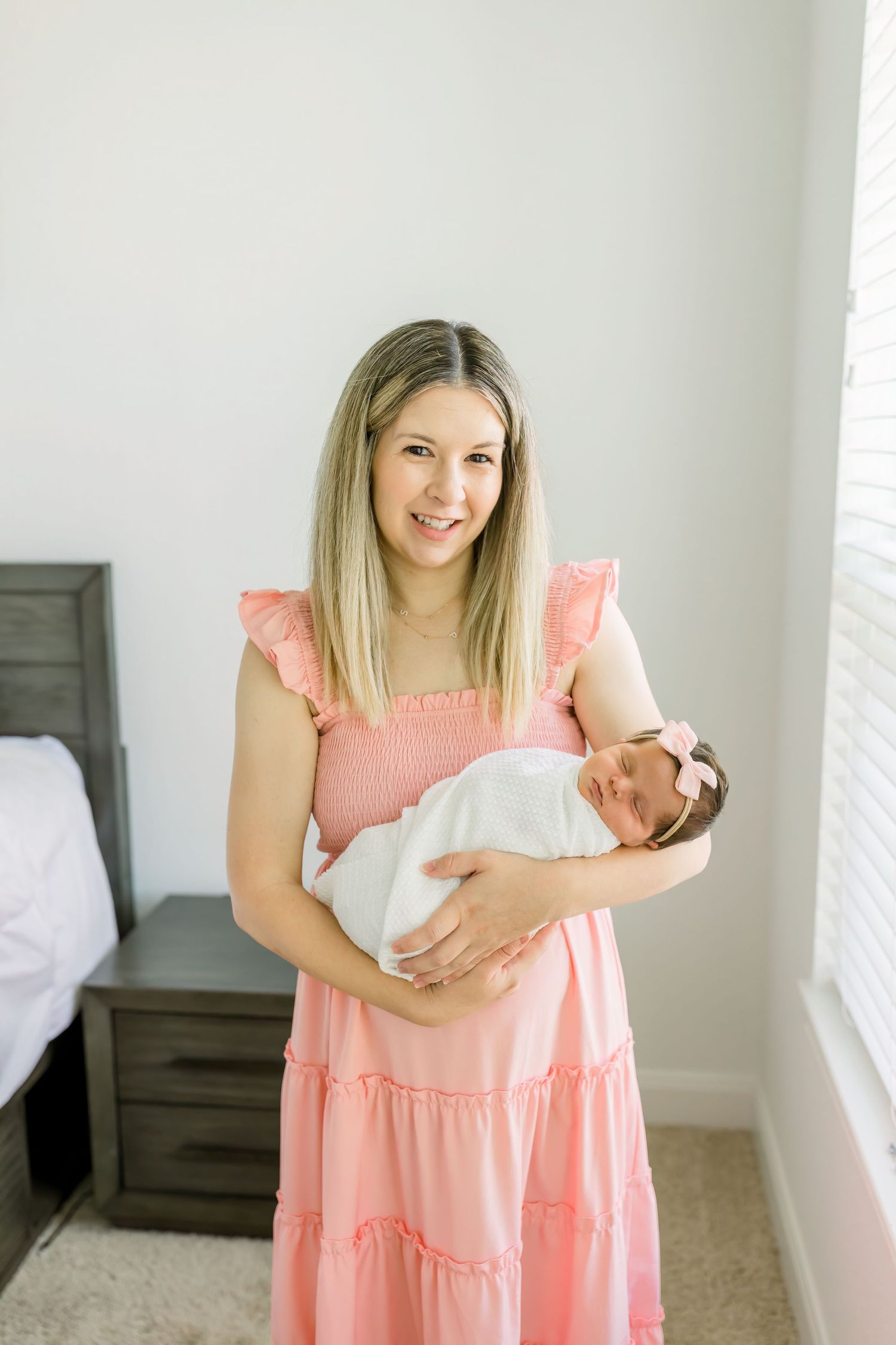 A person in a pink dress holds a baby while standing next to a gray bed.