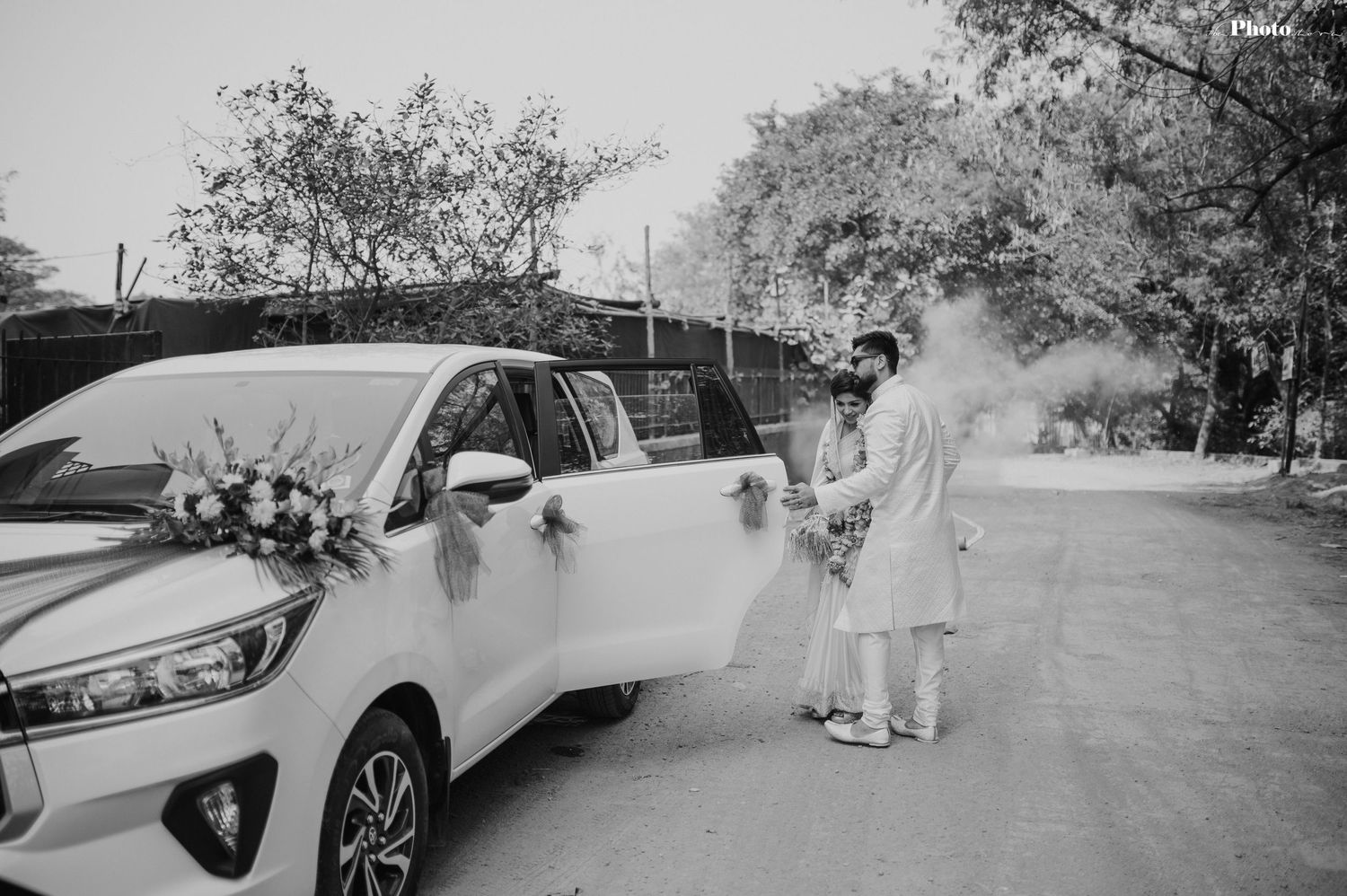 A couple walks hand in hand along a tree-lined pathway in their white wedding outfits.