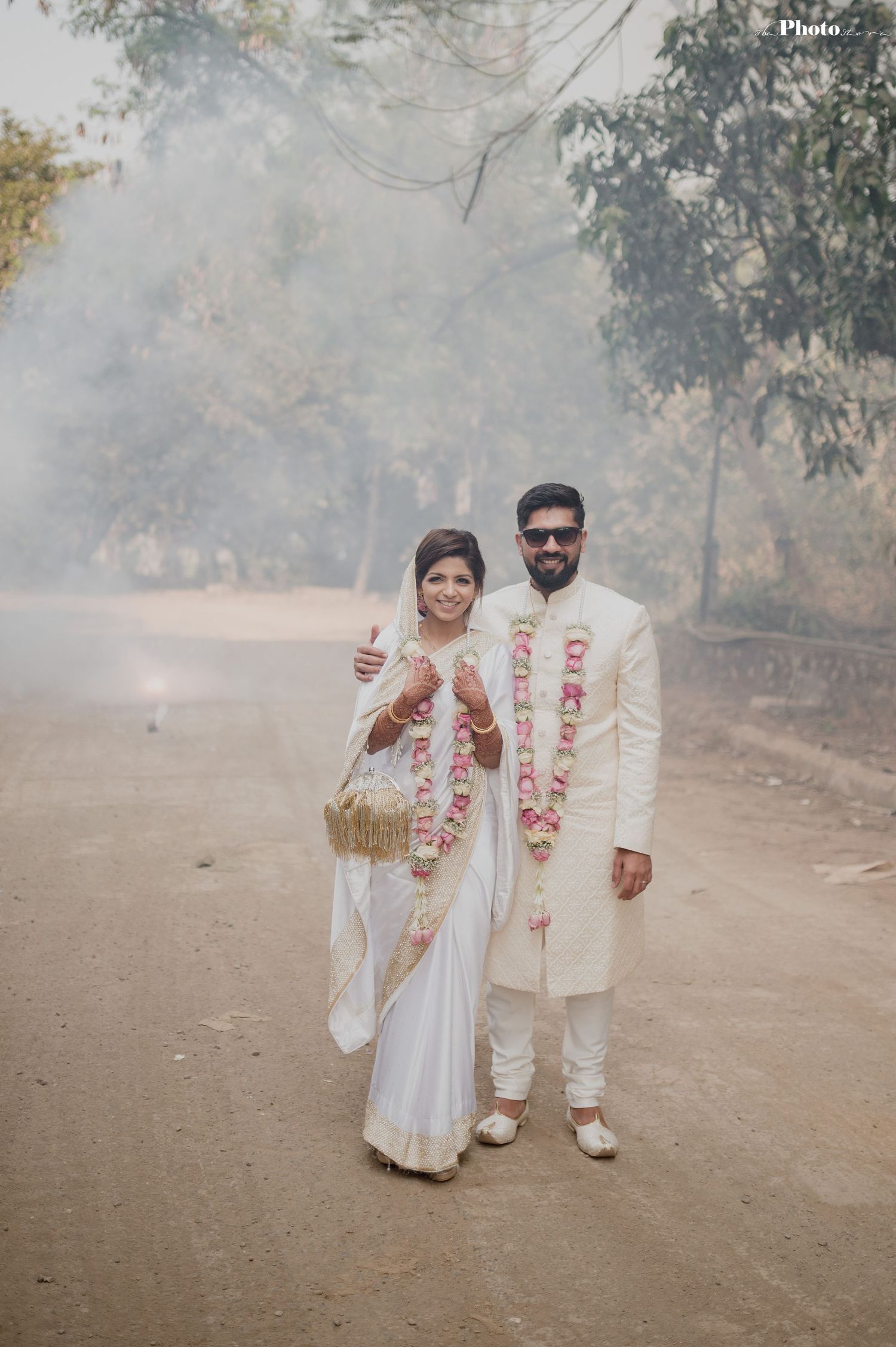 A couple in matching white outfits with pink floral garlands poses on a misty outdoor path.
