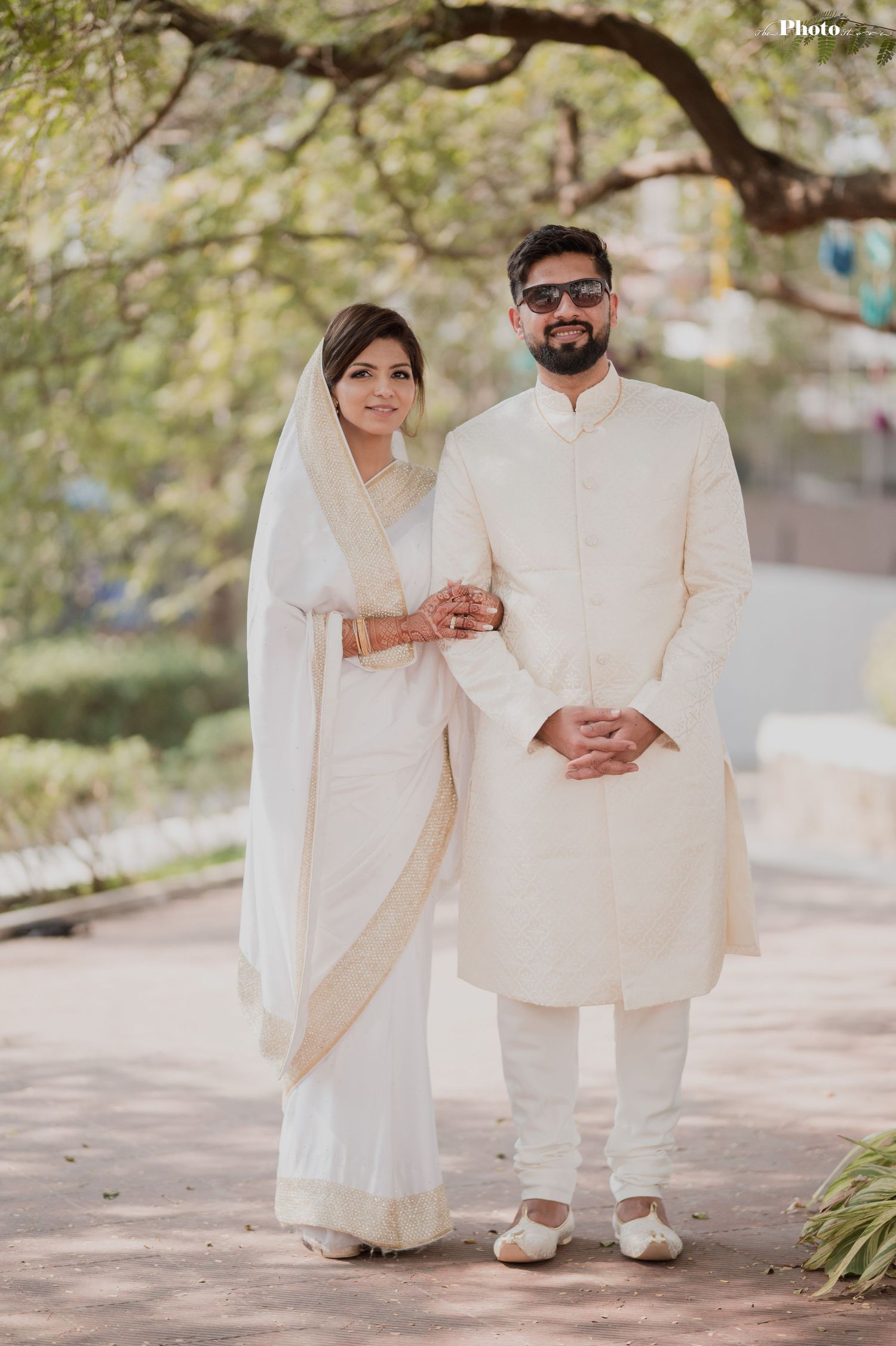 A couple in pristine white wedding attire poses together on a sunlit garden path.