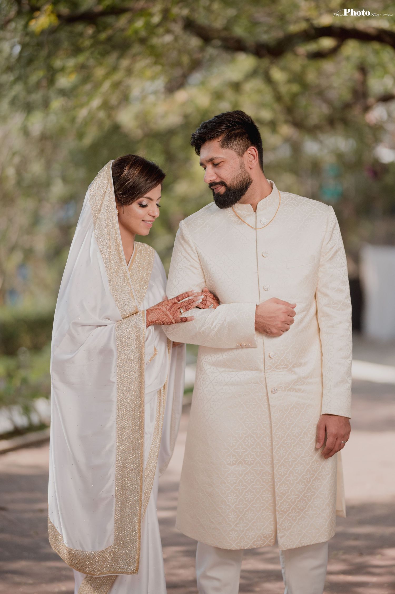 A couple in traditional white wedding outfits shares intimate moments on a tree-lined path.
