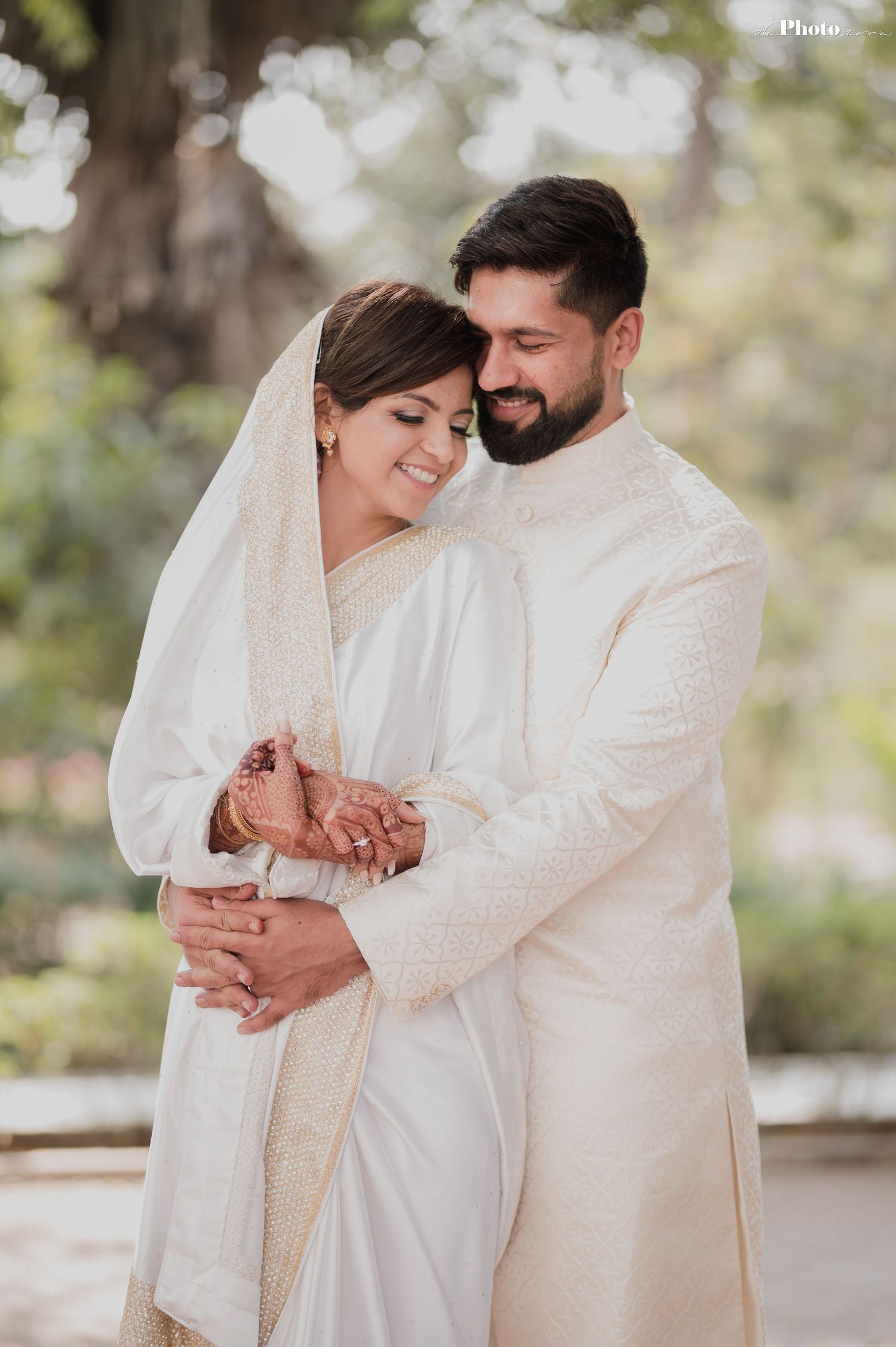 A romantic wedding couple in traditional white outfits embrace tenderly in a sunlit outdoor setting.